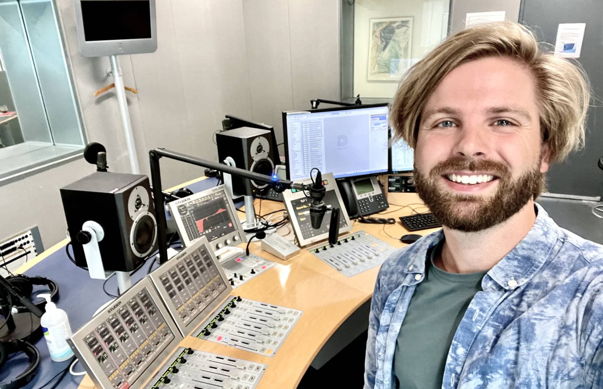 A selfie photo of Nikolaj Stausbøl in the danish public service broadcasting studio. He's facing the back to the studio setup containing knobs and dials and microphones